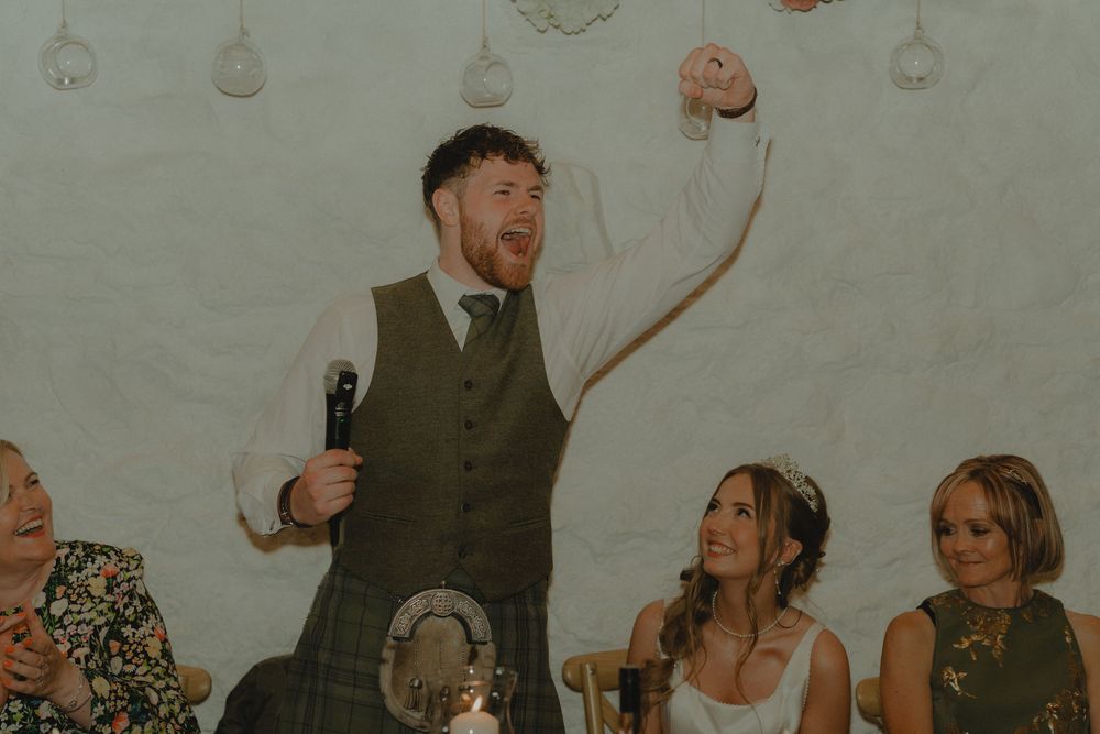 Groom in kilt and waistcoat raises fist during wedding speech while bride and guests smile and clap.