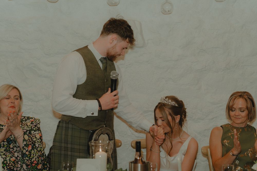Groom in kilt delivers heartfelt wedding speech holding microphone while bride listens emotionally at reception.