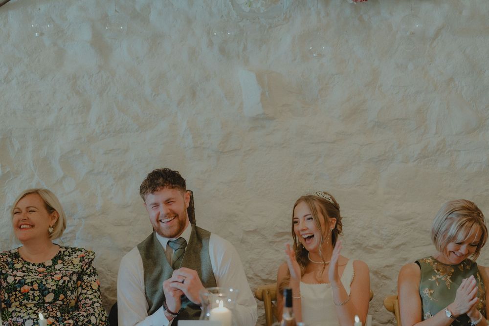 Wedding party laughs joyfully at reception table against rustic white stone wall with warm candlelight.