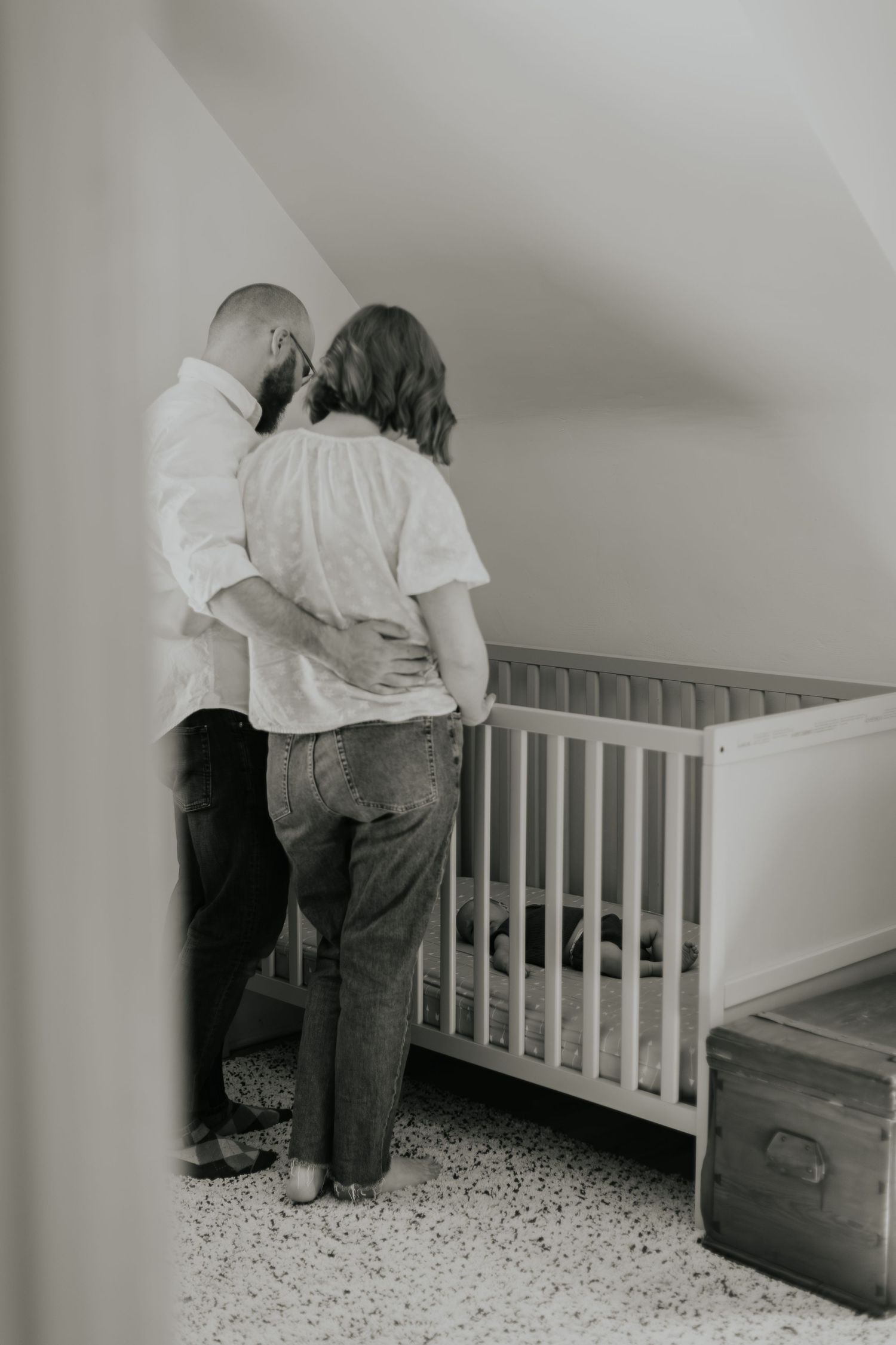 A couple stands together looking into a white crib in a nursery room.