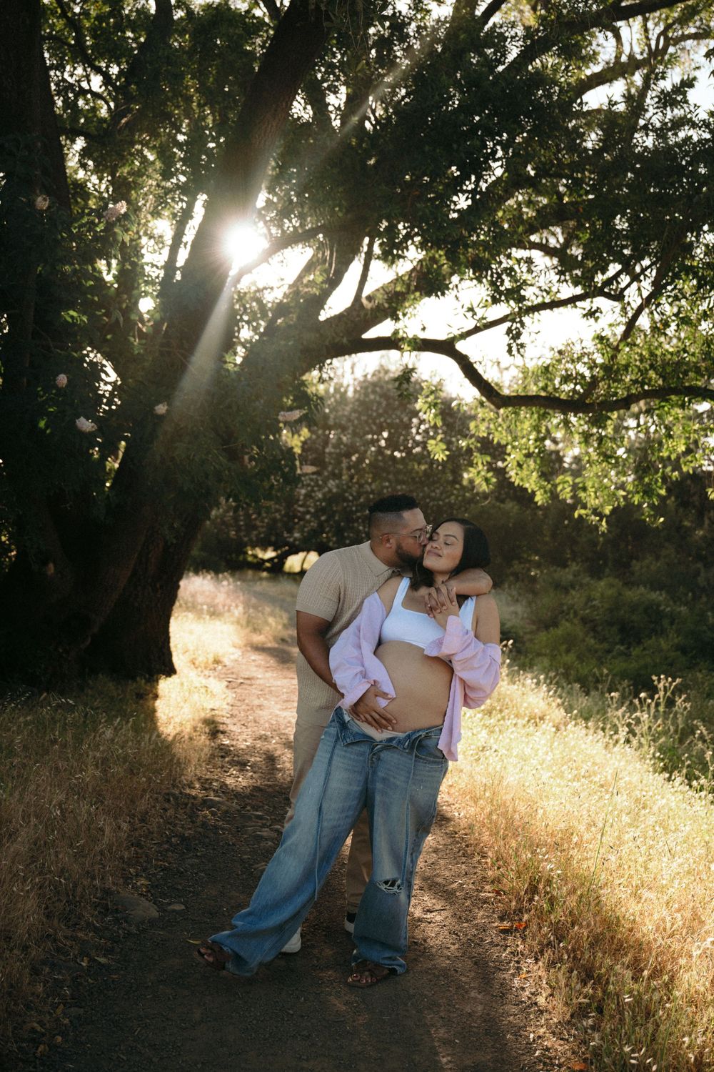 Couple sharing a romantic moment on a sunlit trail beneath a large tree during sunset.