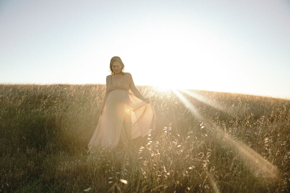 A figure in a flowing white dress stands in a sunlit meadow during golden hour as rays shine through tall grass.