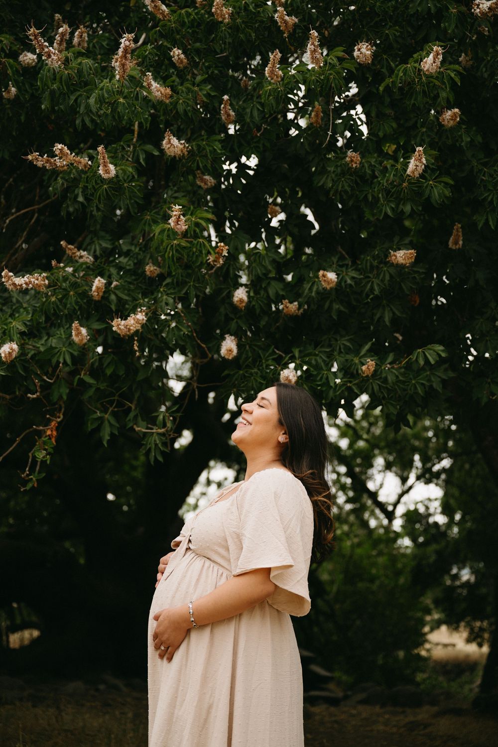 A figure in a flowing white dress poses beneath hanging dried flowers against a dark natural backdrop.