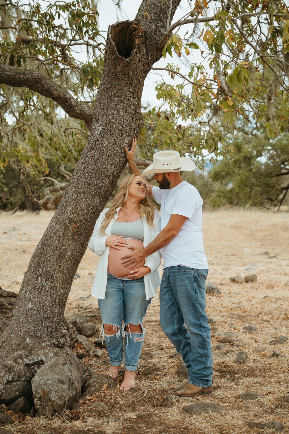 Pregnant couple sharing a tender moment by a tree outdoors, man in cowboy hat cradling woman's baby bump.