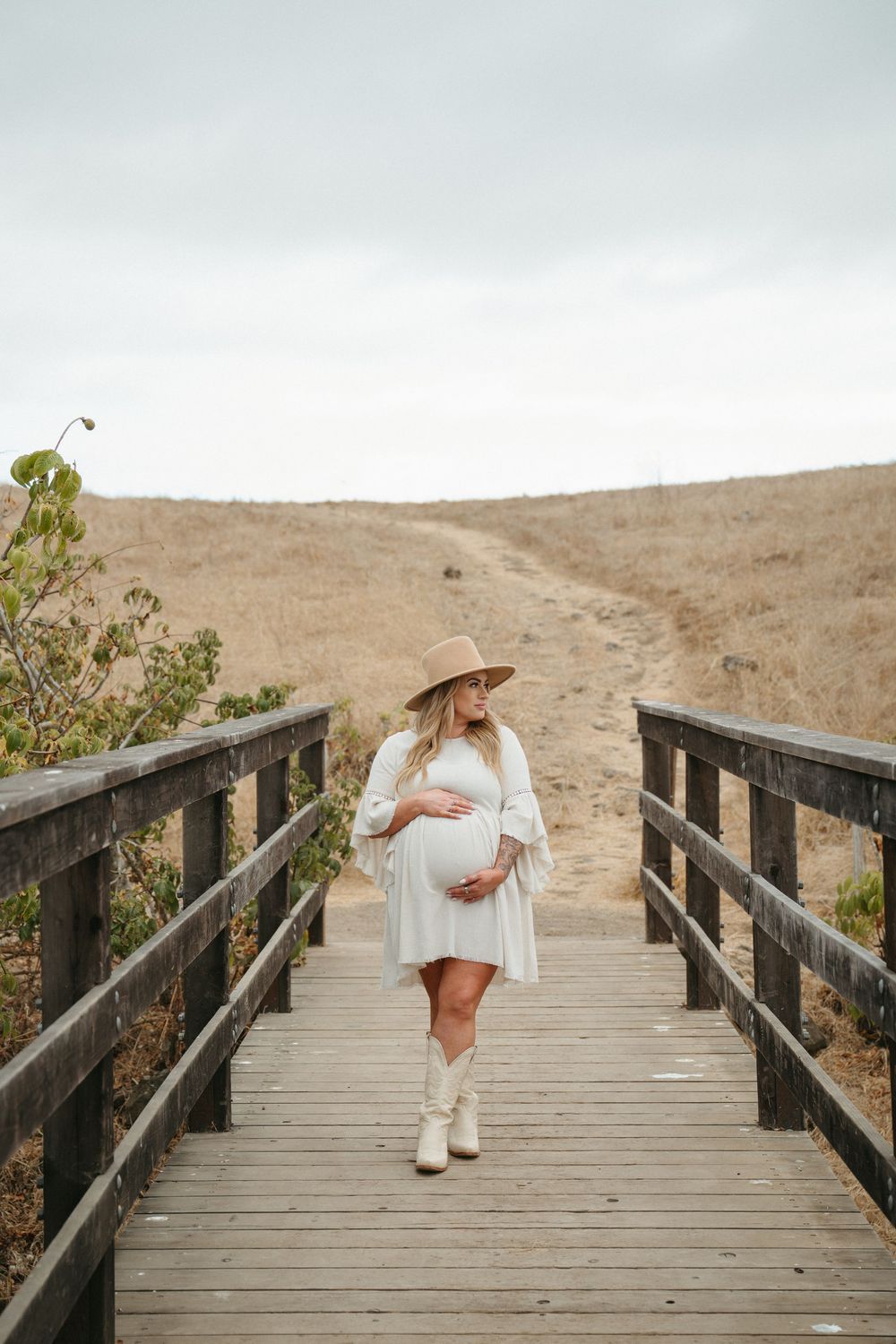 Pregnant woman in white dress, tan hat, and cowboy boots cradling her belly on a wooden boardwalk path.
