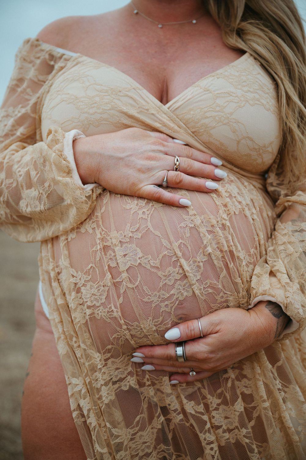 Pregnant woman in sheer beige lace maternity gown cradling her baby bump, wearing silver rings, outdoor beach setting.