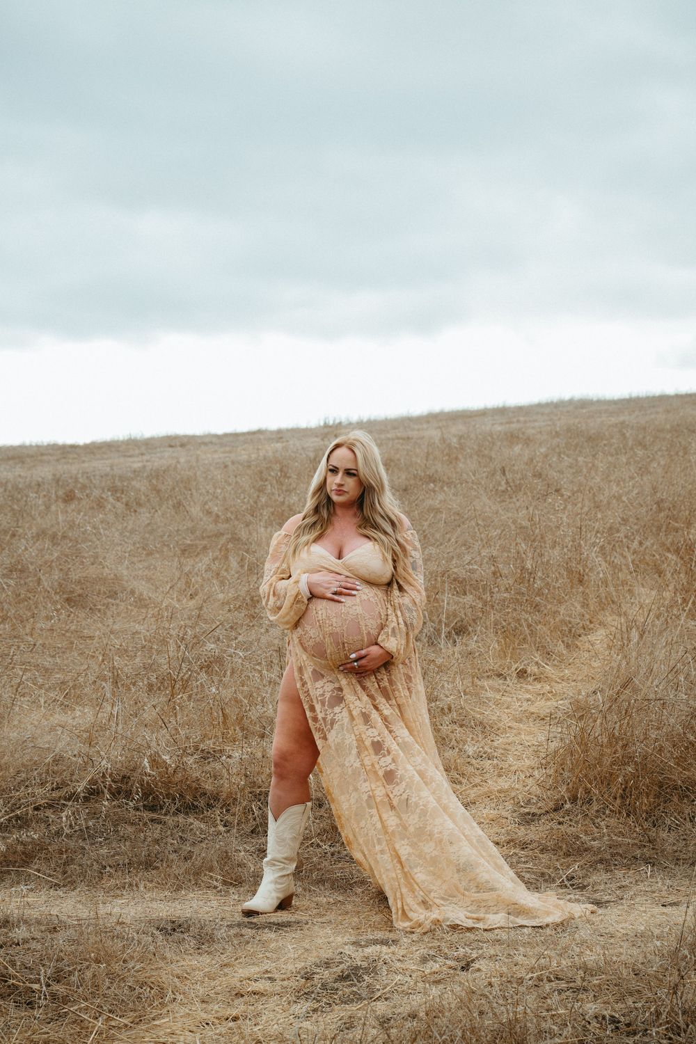 Pregnant woman in sheer floral gown and white boots standing in golden field under cloudy sky.