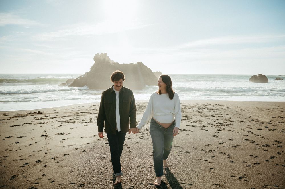 Couple holding hands walking on a sunny beach with ocean waves and rocky cliffs in the background.