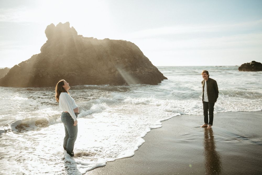 Couple standing on a sunlit beach with ocean waves and dramatic rock formations in the background.