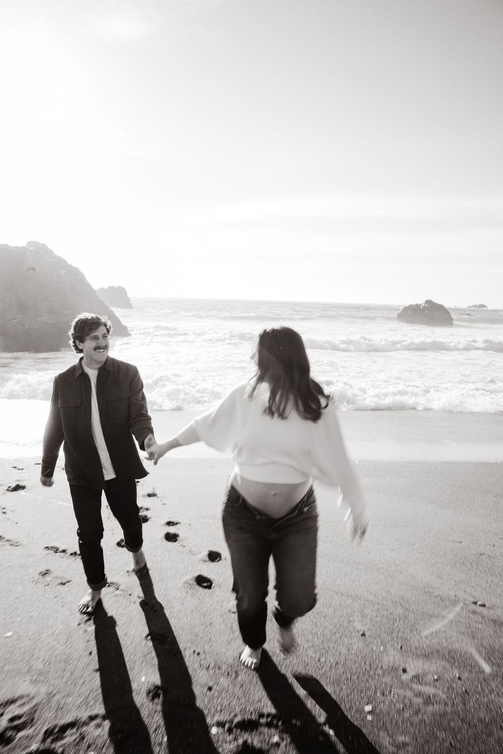 Pregnant couple holding hands and walking barefoot on a black sand beach in black and white photo.