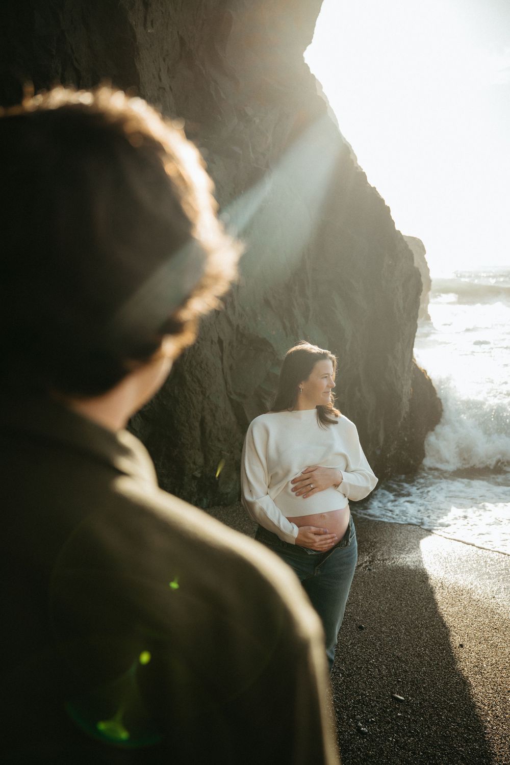 Pregnant woman cradling baby bump on rocky beach with sunlight streaming through cliffs while partner watches.