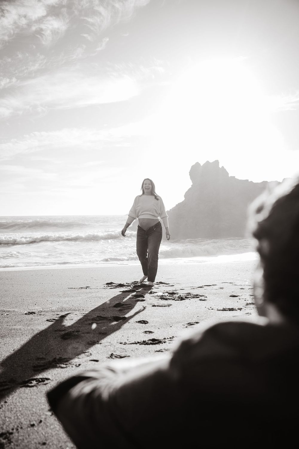 Black and white photo of a woman walking on a sunny beach toward the camera, with ocean waves and rocky cliffs behind her.
