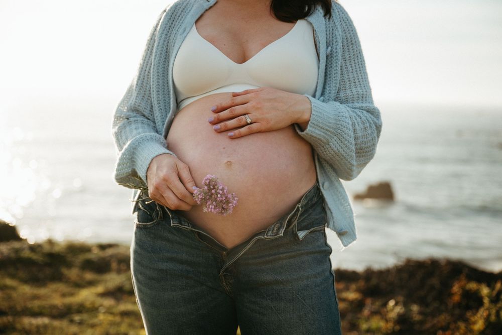 Pregnant woman in blue cardigan and jeans holding a flower against her bare belly near the ocean.