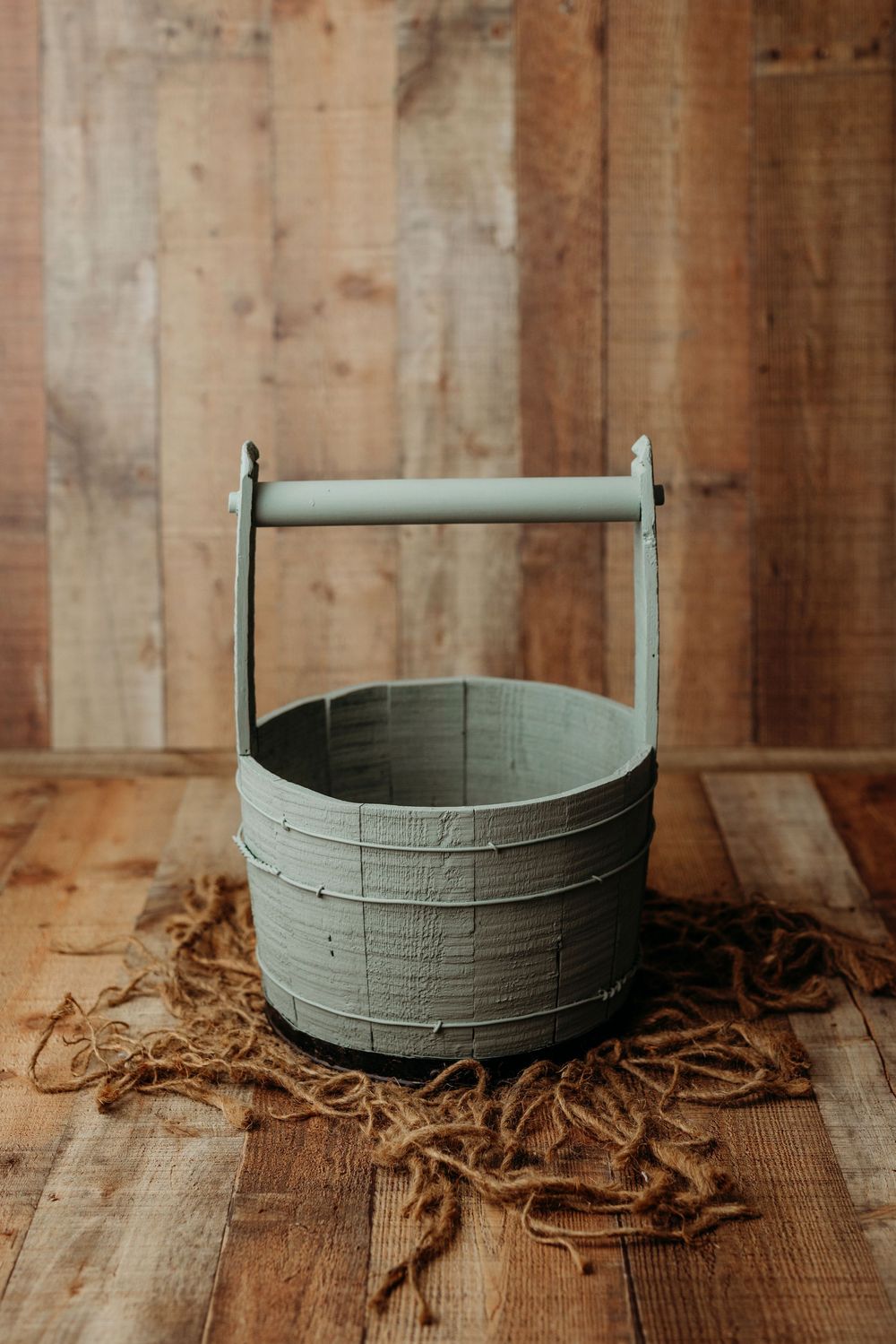 A vintage-style sage green wooden bucket with handle sits on rustic wooden surface with scattered wood shavings.