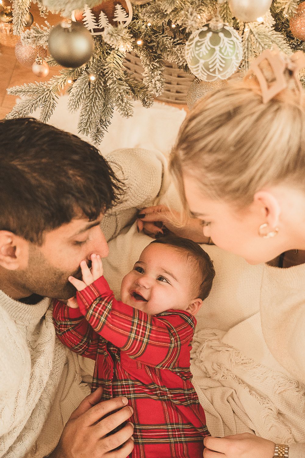 A loving Christmas portrait series under decorated evergreen branches with warm lighting and plaid holiday attire.