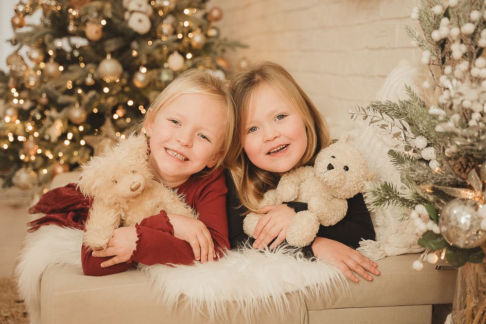 Children lying together on a cozy white blanket in front of a decorated Christmas tree with glowing lights.