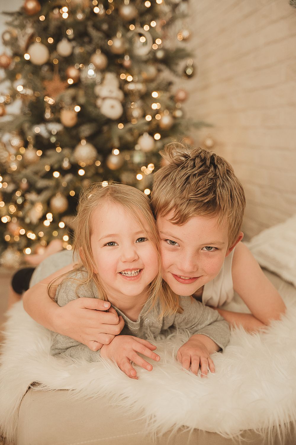 Two kids laying on white furry blanket smiling and laughing together with Christmas tree with lights in background.