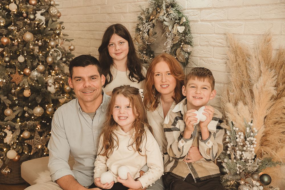 Family sitting together on a couch in front of a decorated Christmas tree in warm neutral tones.