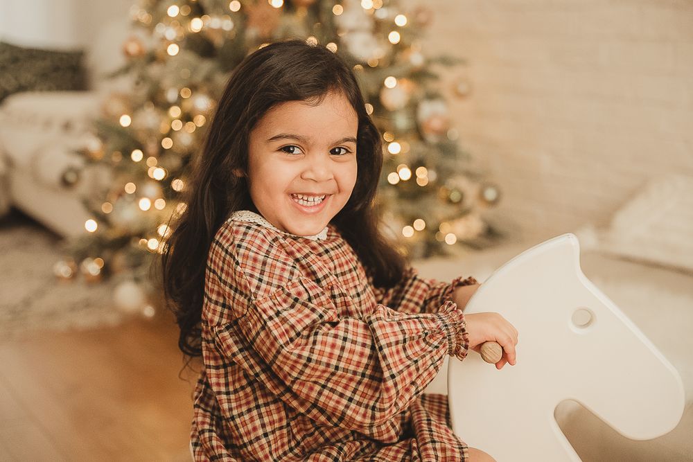 A child in a brown plaid dress poses cheerfully in front of a sparkly Christmas tree with golden lights and decorations.