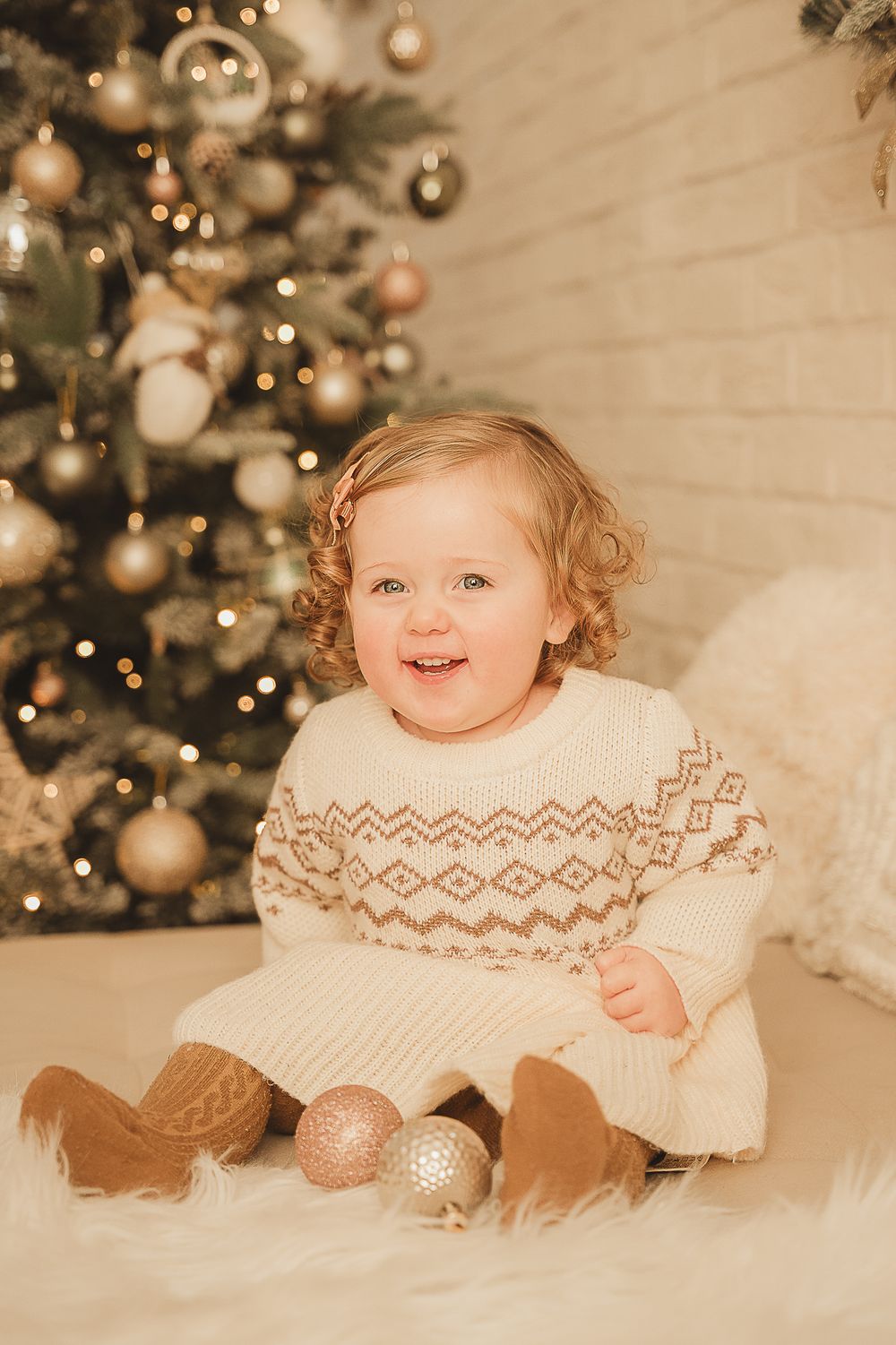 A small child in a festive white sweater sits on a white fur rug with Christmas ornaments and a decorated tree.