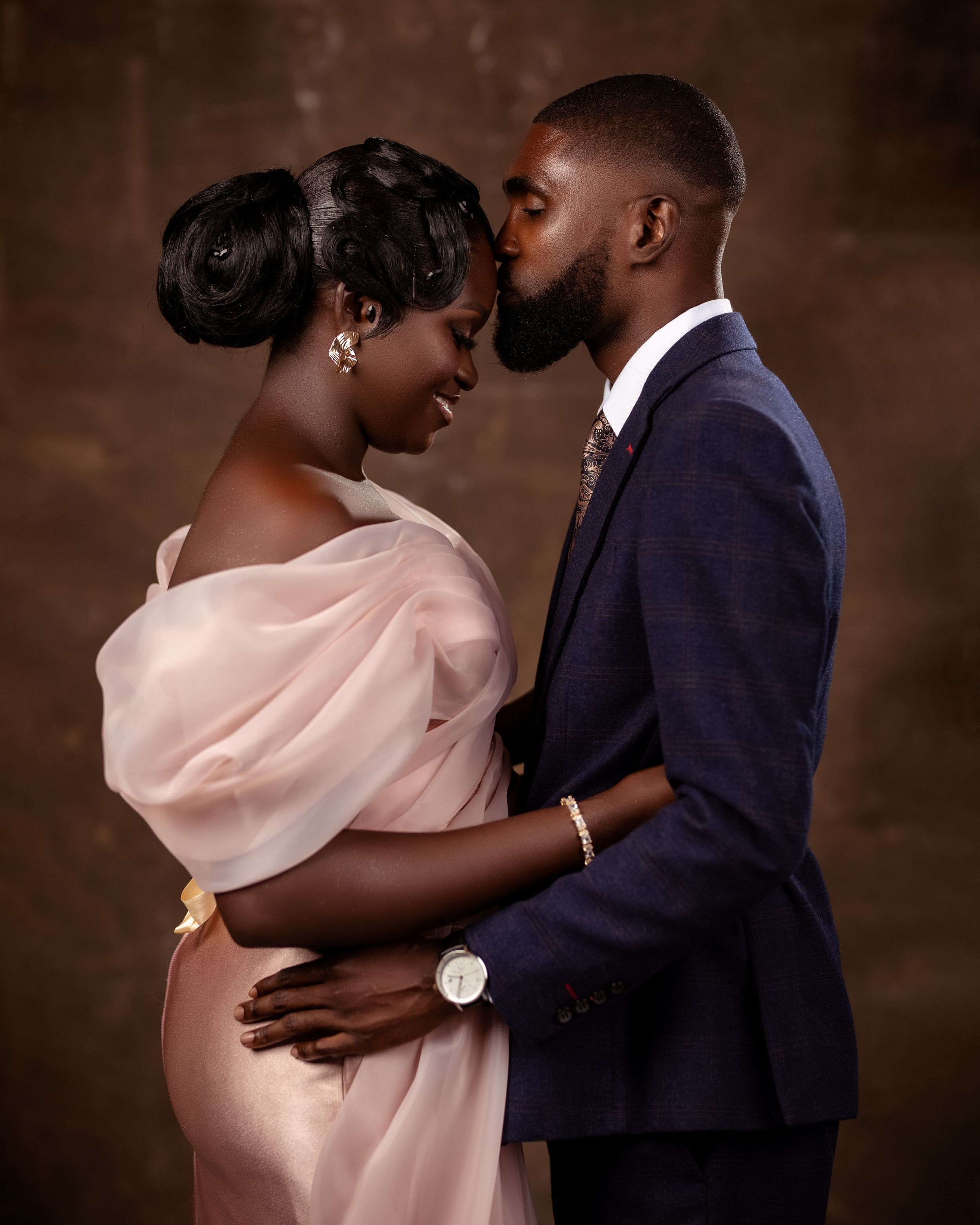 Romantic studio portrait of a couple in formal wedding attire posed against a dark brown background.