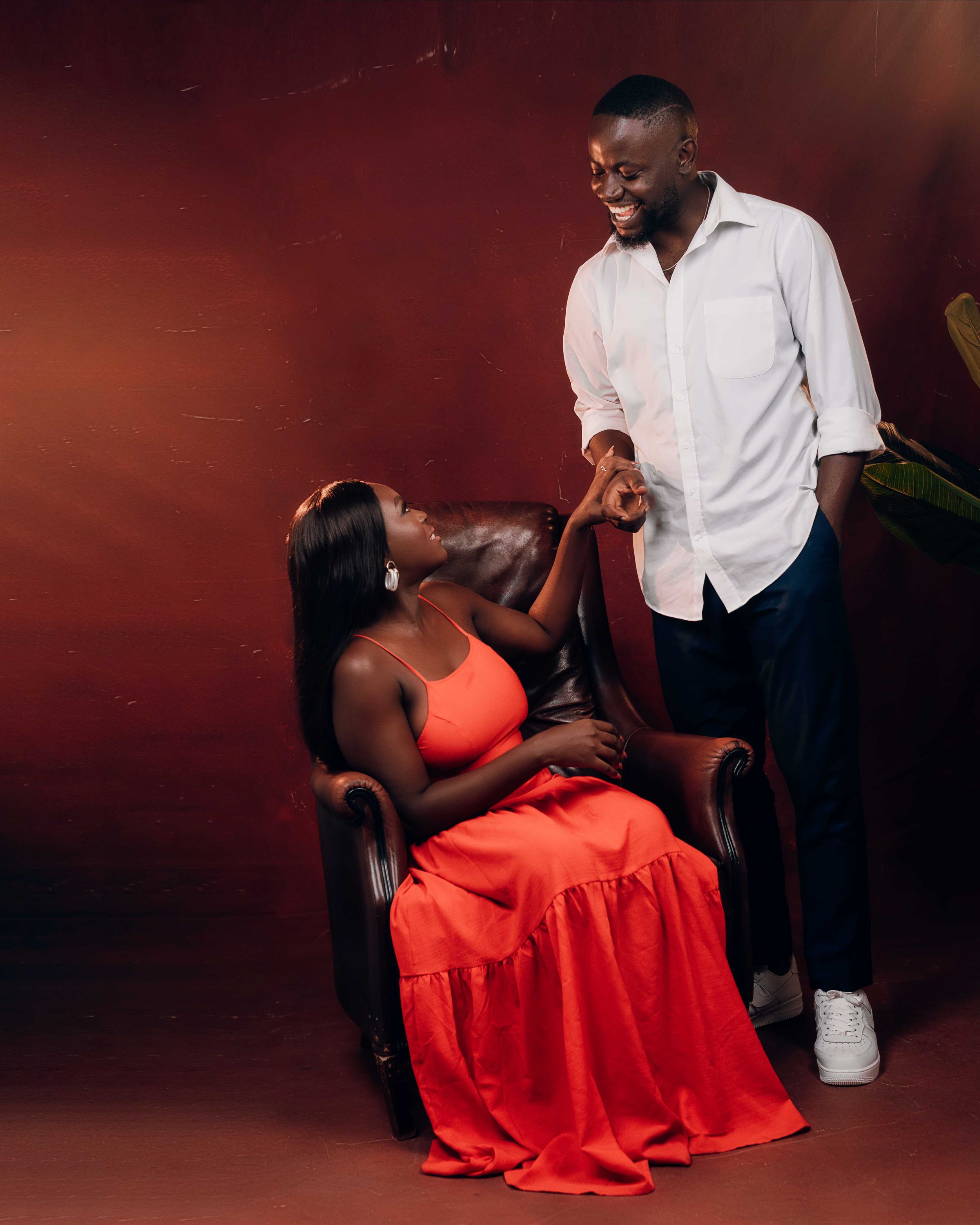 Romantic indoor studio session of a couple, with the woman in a bold red dress and the man in a white button-down, posed in a leather chair