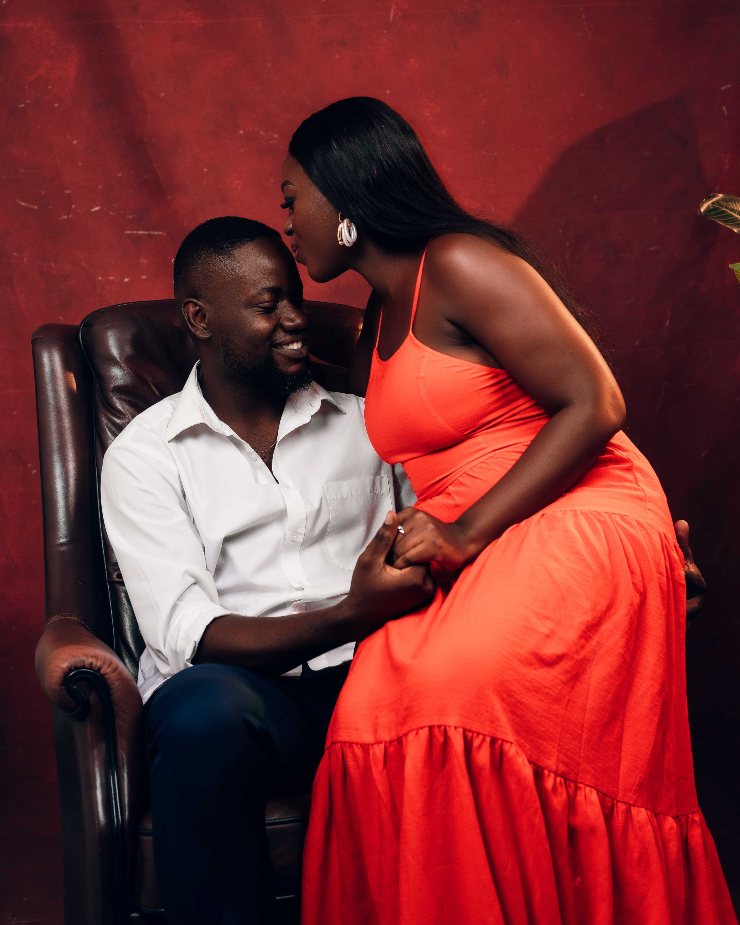 Romantic indoor studio session of a couple, with the woman in a bold red dress and the man in a white button-down, posed in a leather chair