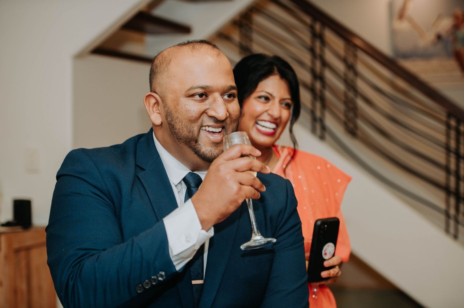Two guests enjoy drinks and laughter at an elegant indoor celebration.