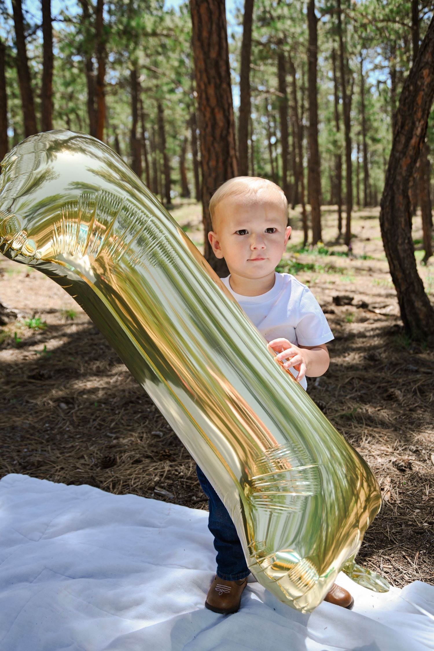 Little one stands outdoors in a pine forest holding a large reflective gold mylar balloon on a white blanket.