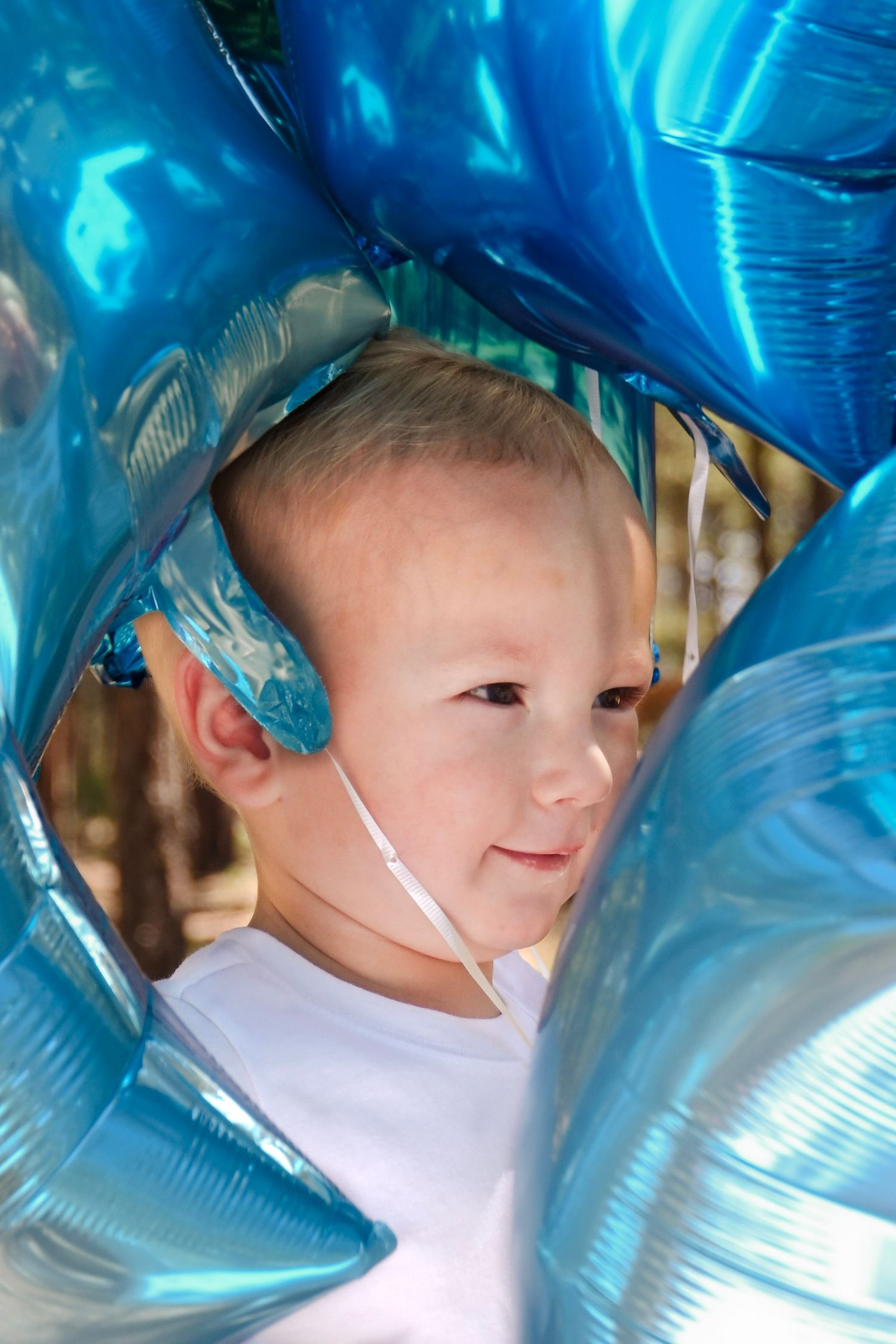 A child peers through blue metallic balloons wearing a white t-shirt as they smile softly.