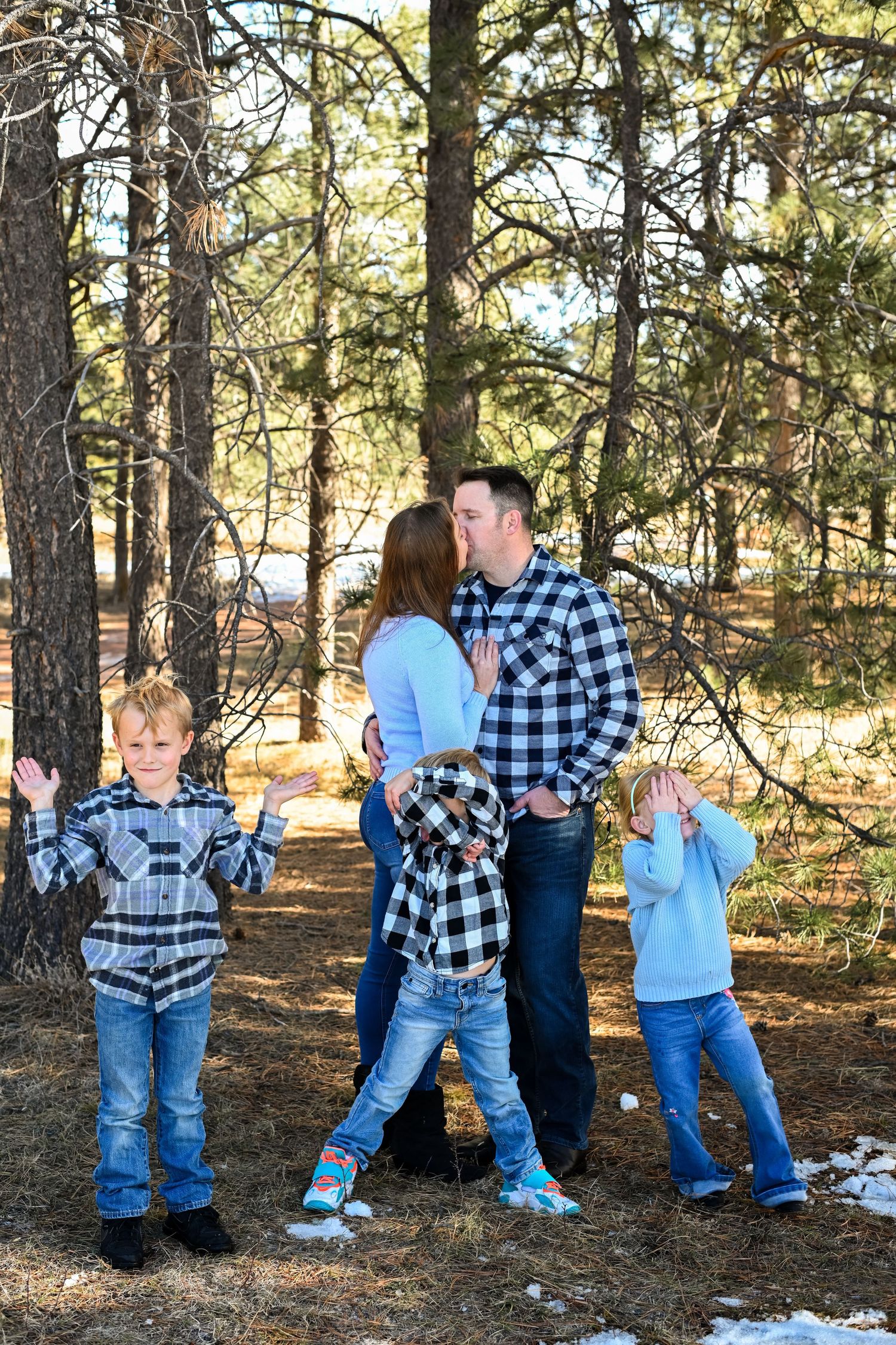 A family wearing coordinated blue plaid shirts poses together in a forest setting during winter.