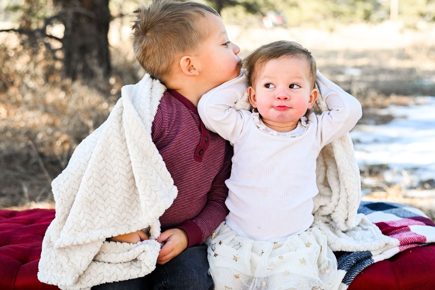Two children bundled in white blankets sit together on a red picnic blanket during a winter outdoor photo session.