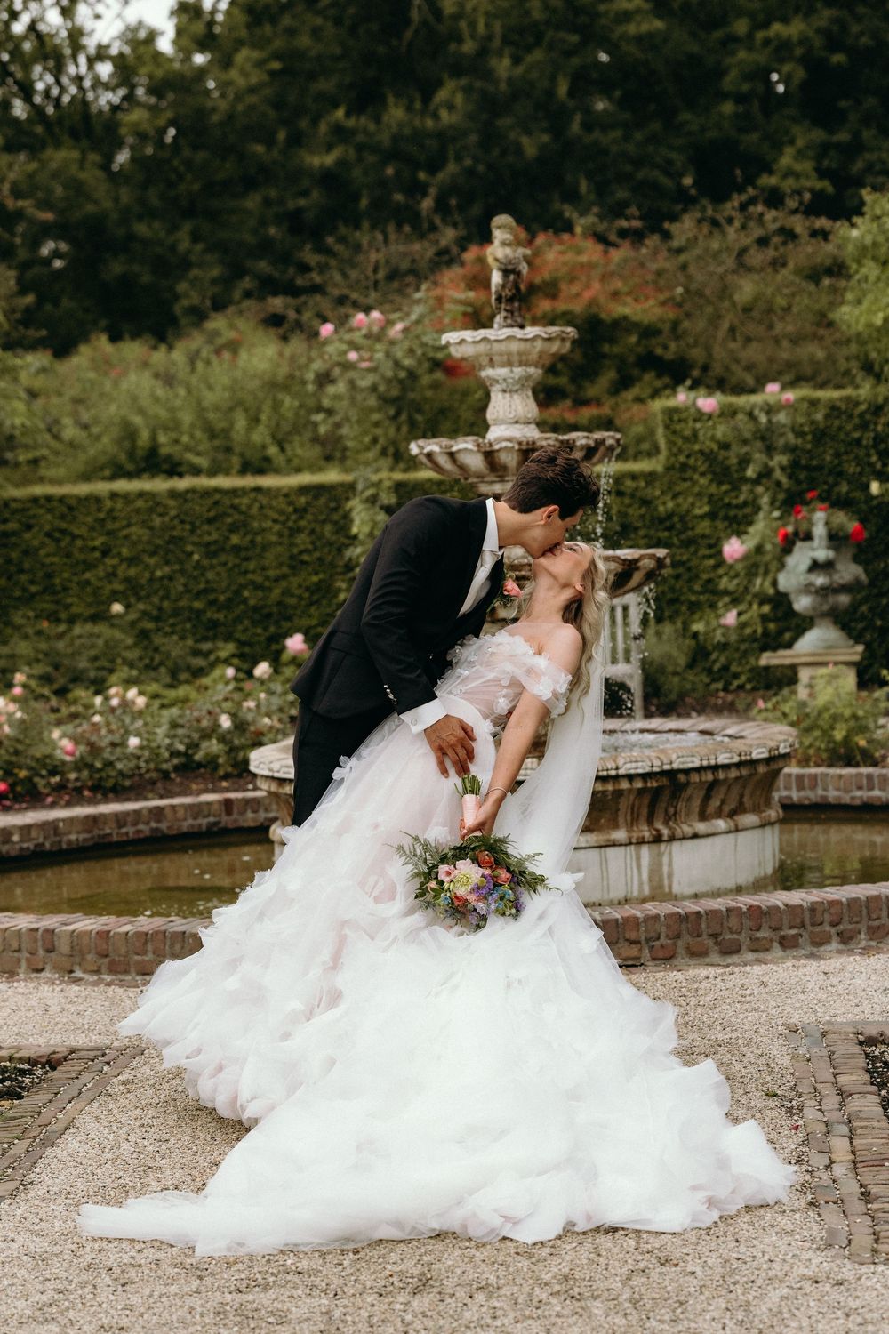 Romantic wedding portrait by an ornate fountain in a formal garden setting.