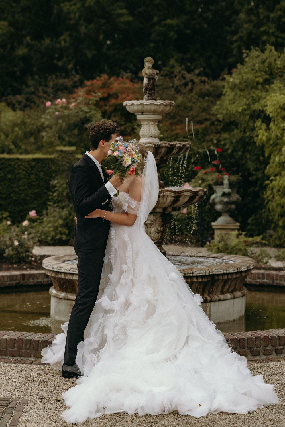 Elegant wedding couple embracing near a classical stone fountain in a manicured garden.