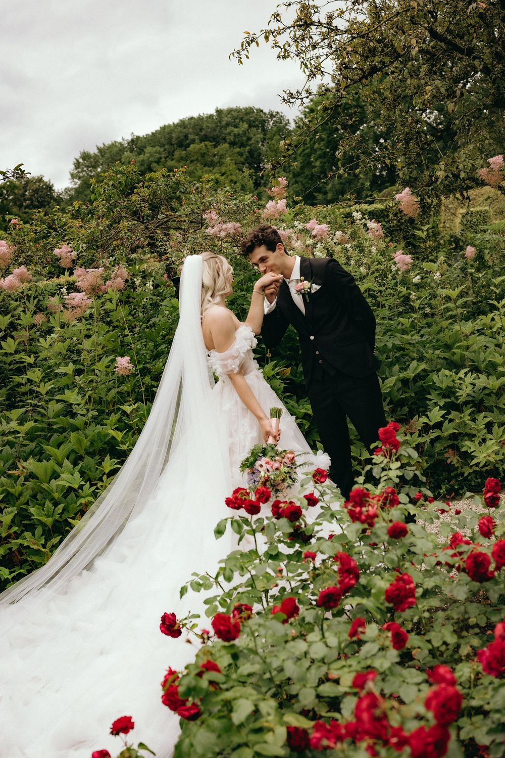 Wedding couple surrounded by vibrant red rose bushes in a lush garden setting.