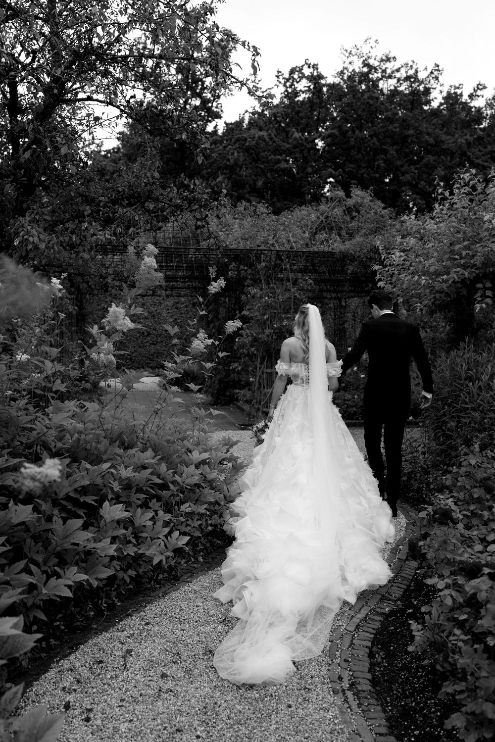 A flowing white wedding dress trails along a garden path surrounded by lush greenery.