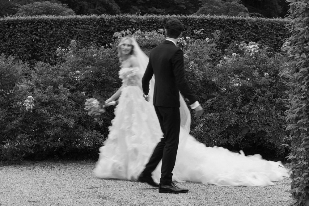 A wedding couple walks along a gravel path with trees and foliage in the background.
