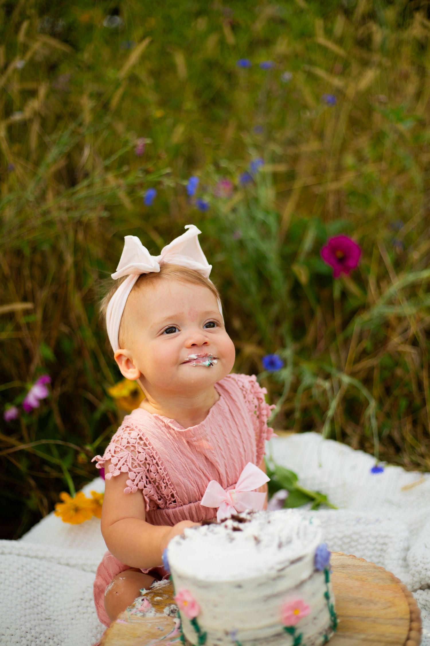 Baby in pink dress with bow headband enjoys first birthday cake smash photo session.