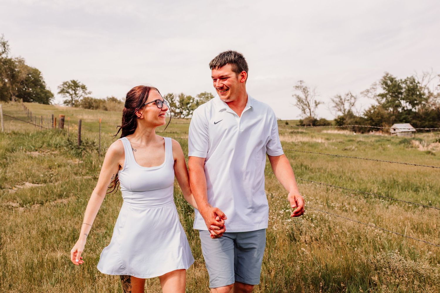 A young couple holds hands and smiles while walking through a lush green field on a sunny day.