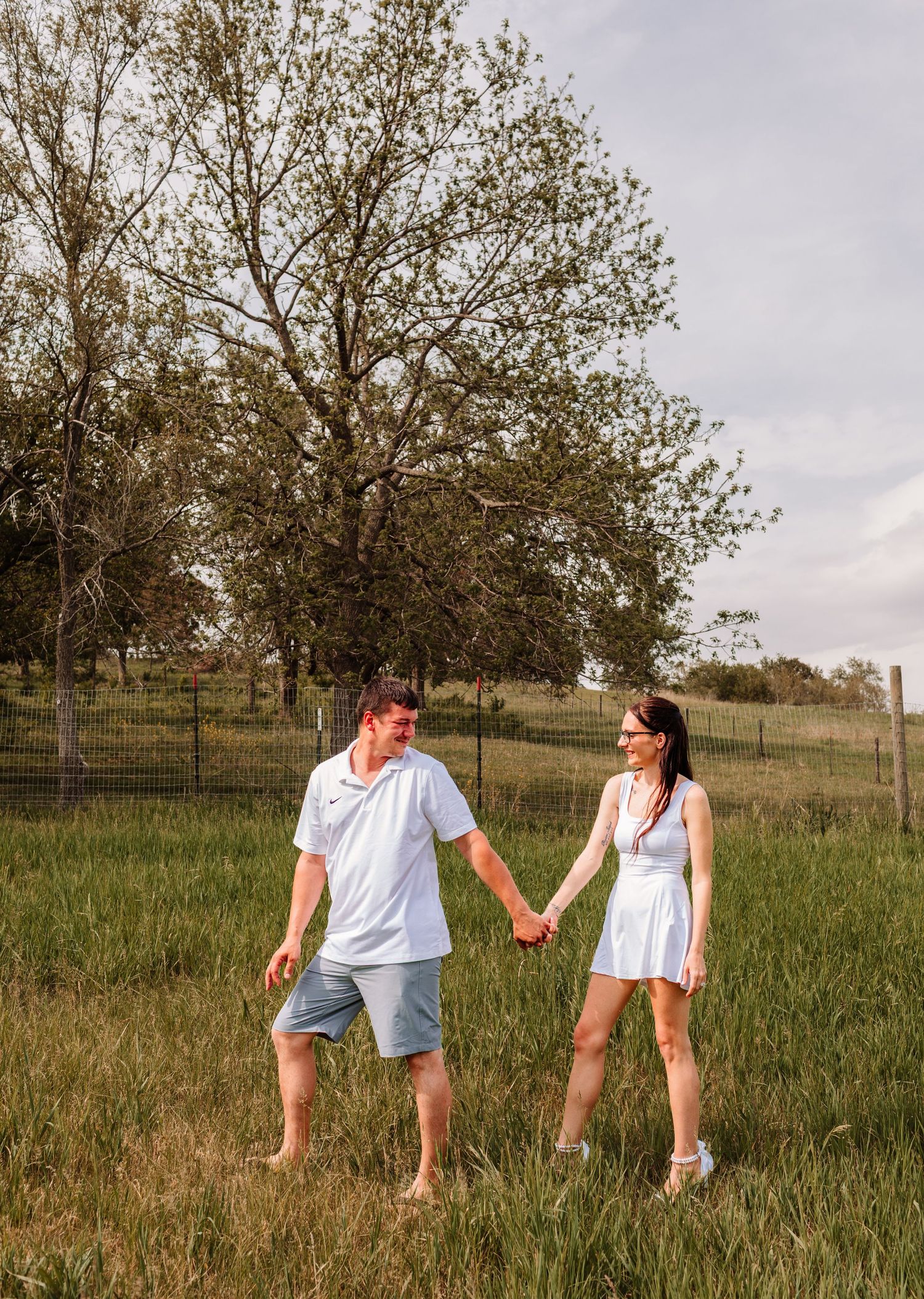 Couple holding hands walking through green field with trees in background on sunny day.