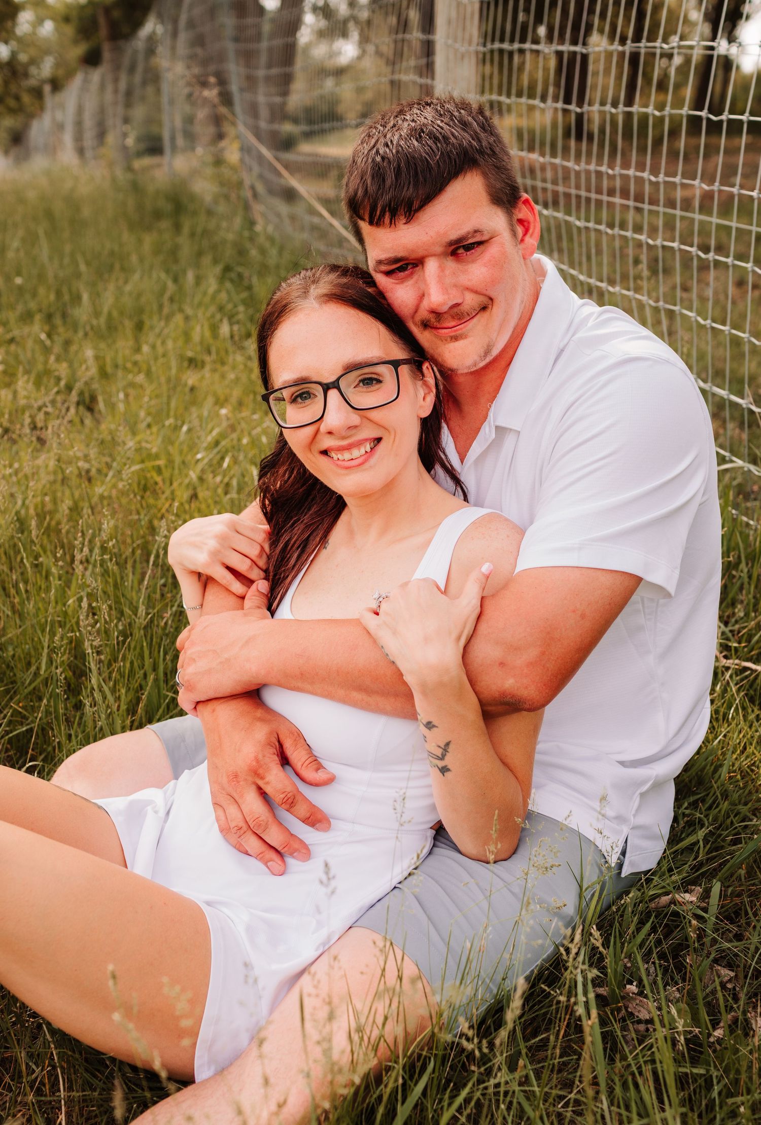 Smiling couple sitting in grass leaning against fence, woman wearing glasses in white dress.