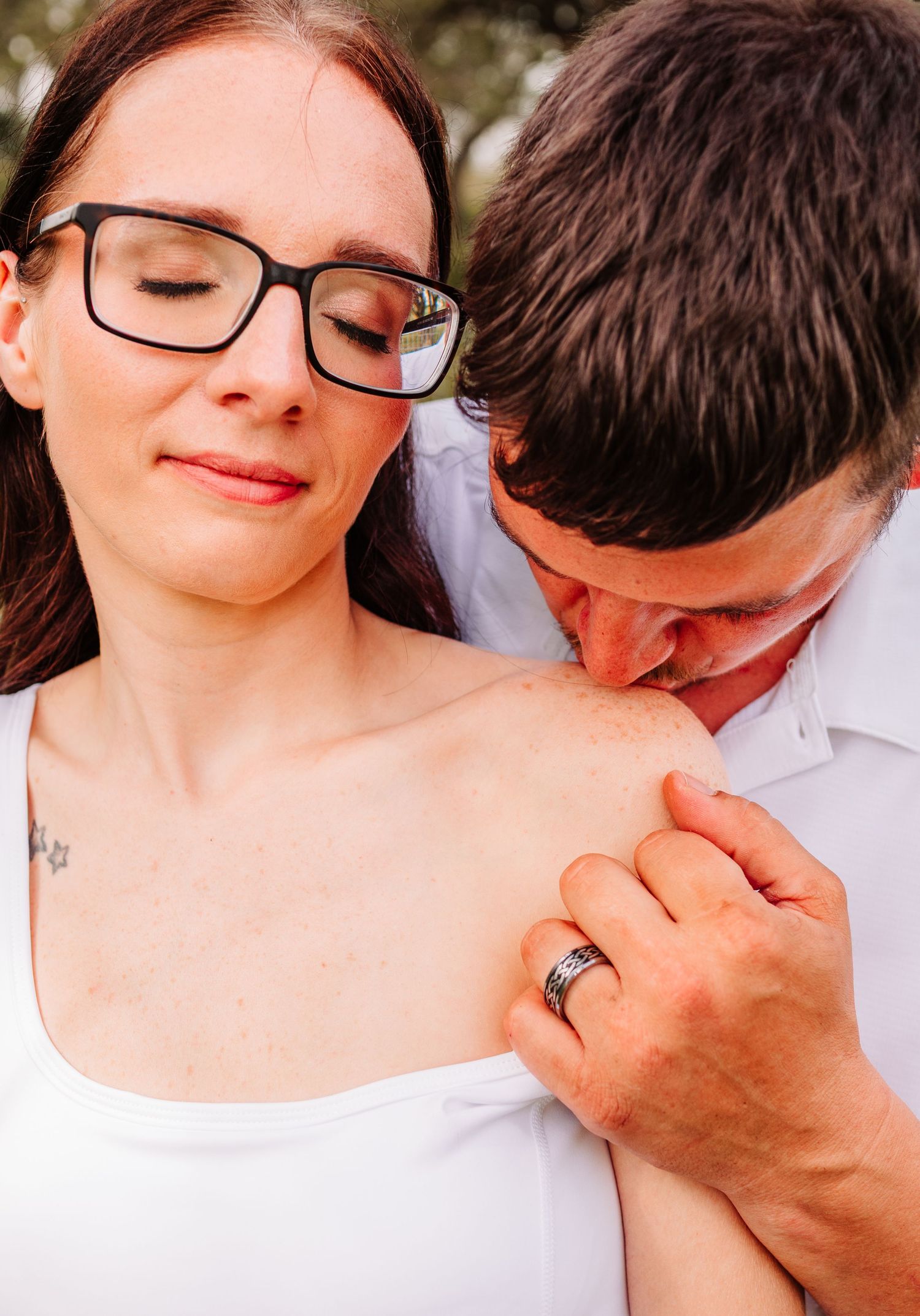 Romantic couple embracing outdoors, woman with black glasses and tattoo, man kissing her shoulder wearing a wedding band.