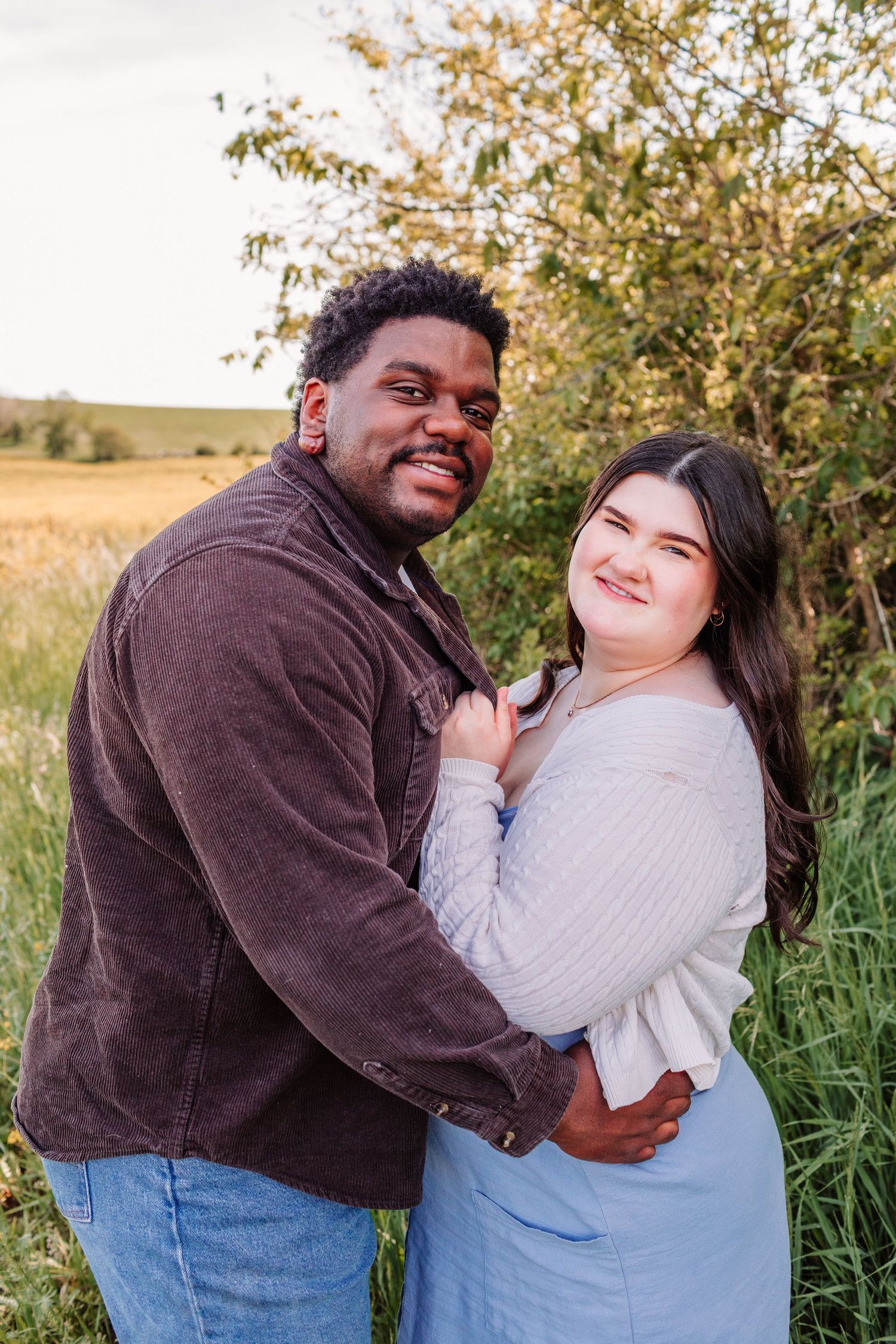 A smiling couple embraces outdoors in a green field, the man in a brown jacket and the woman in a light blue skirt.