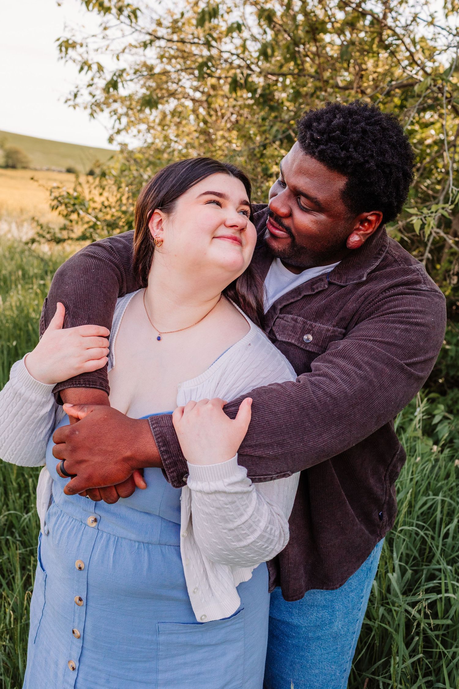 A happy interracial couple embracing outdoors in a green field, the man hugging the woman from behind as she smiles up at him.