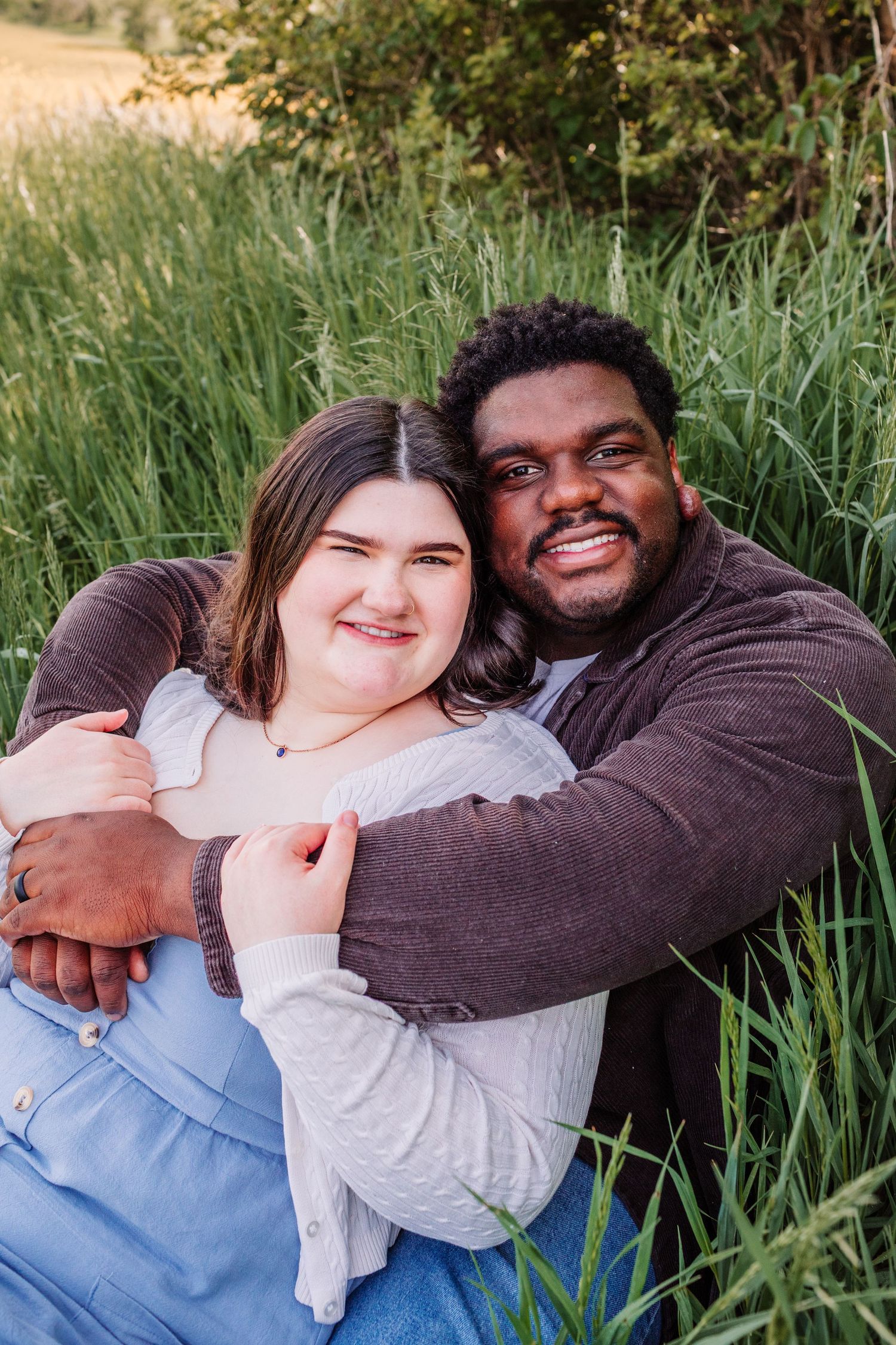 A happy couple embraces while sitting in tall green grass, smiling warmly at the camera in a sunlit outdoor setting.