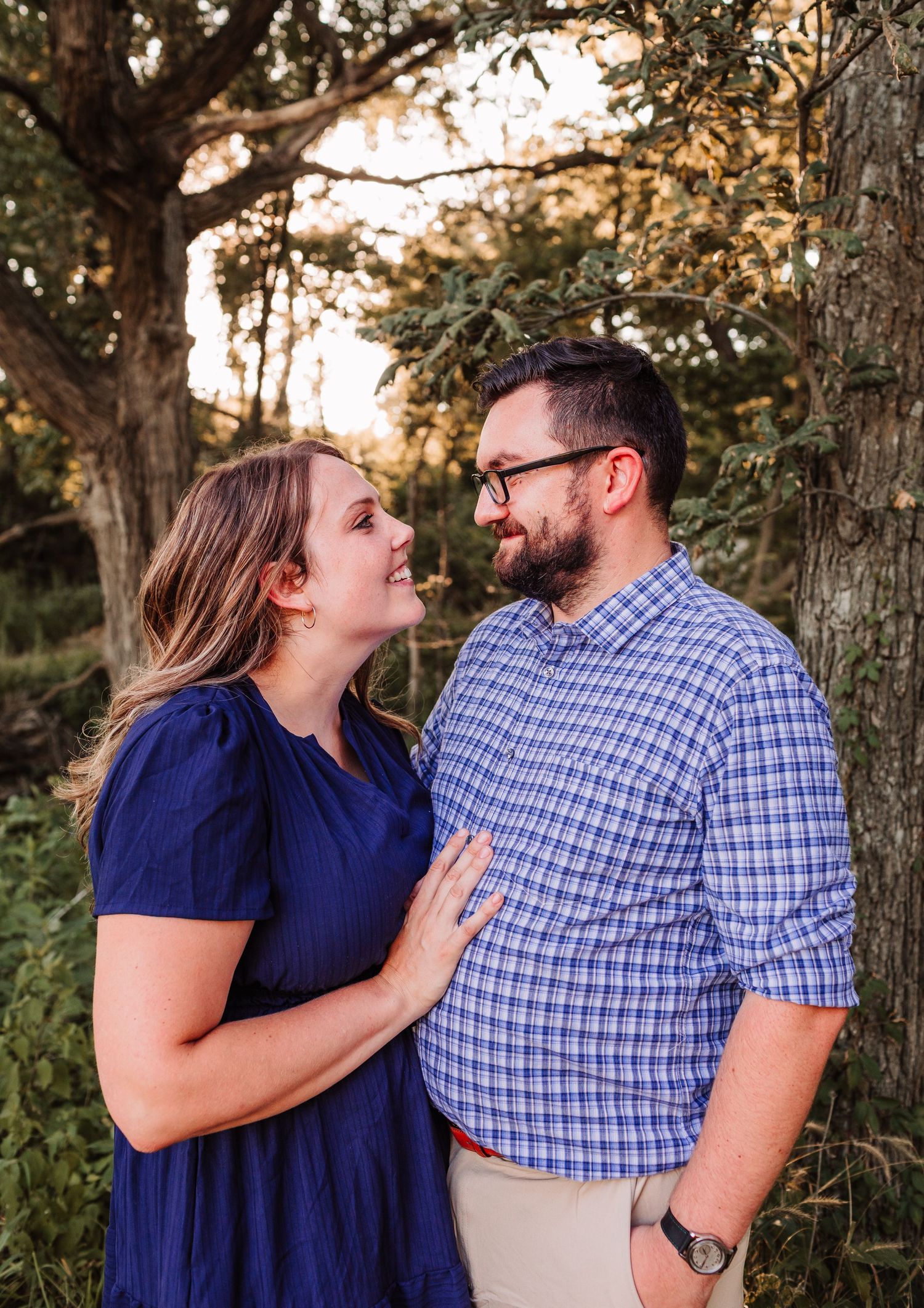 A couple gazes at each other lovingly outdoors, woman in navy dress, man in blue plaid shirt, golden hour lighting.