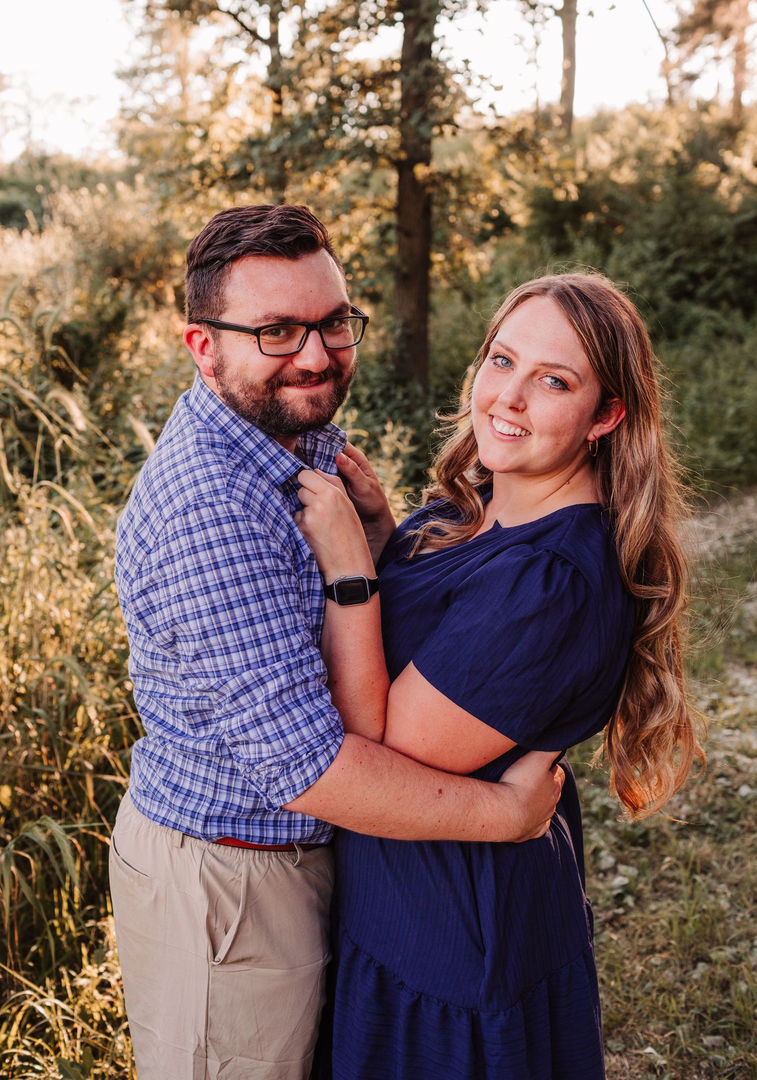 A smiling couple embraces outdoors at golden hour, man in plaid shirt with glasses, woman in navy dress, surrounded by greenery.