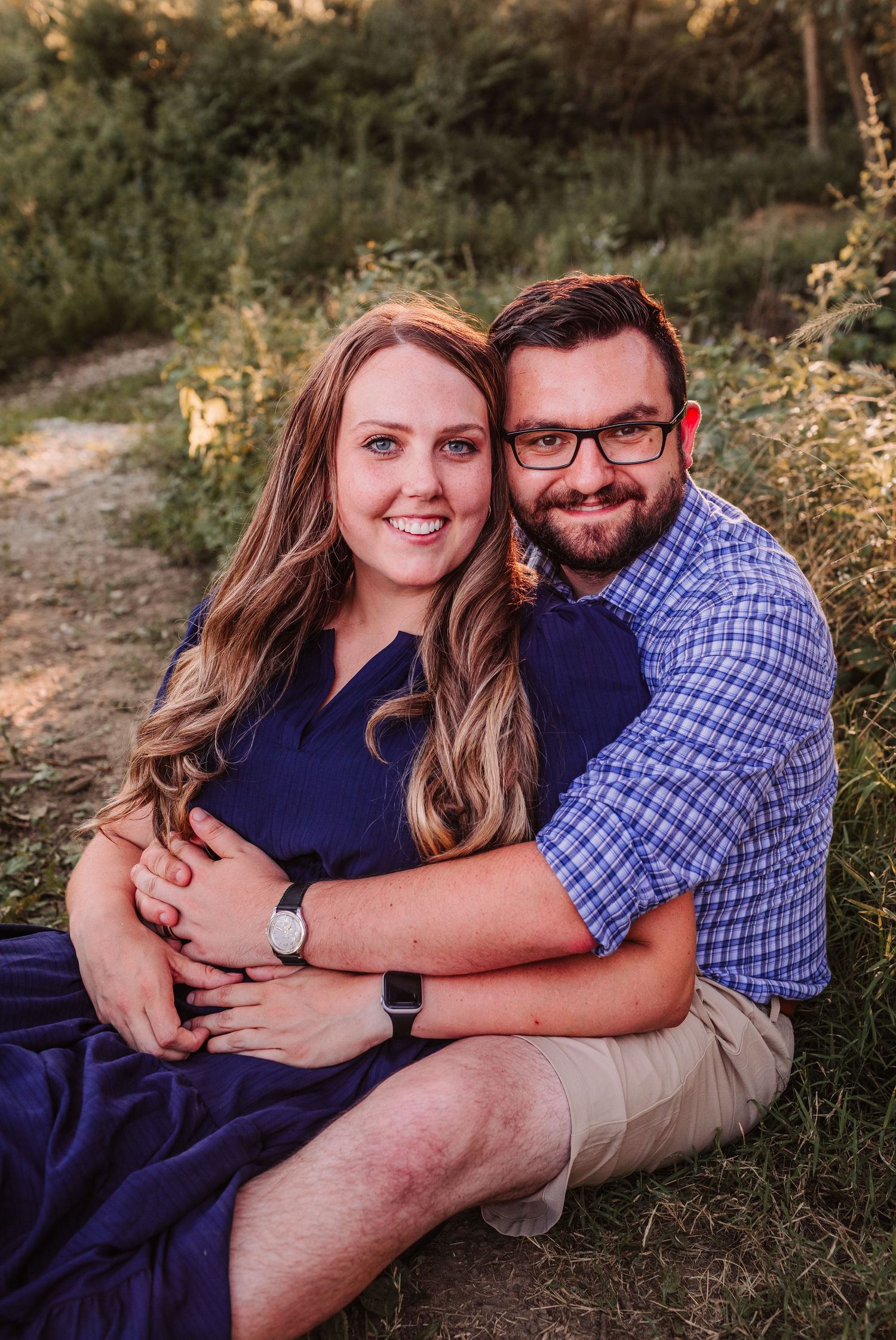 A smiling couple sits outdoors on grass, the man embracing the woman in a navy dress, with lush greenery behind them.