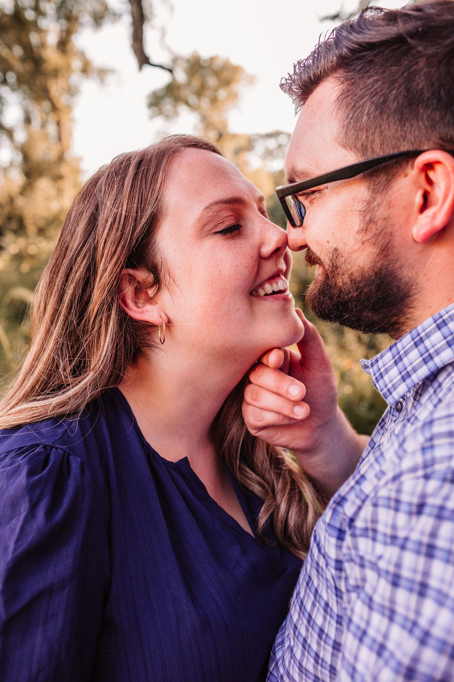 A smiling couple sharing a tender nose-to-nose moment outdoors, the man in glasses and the woman in a blue top.