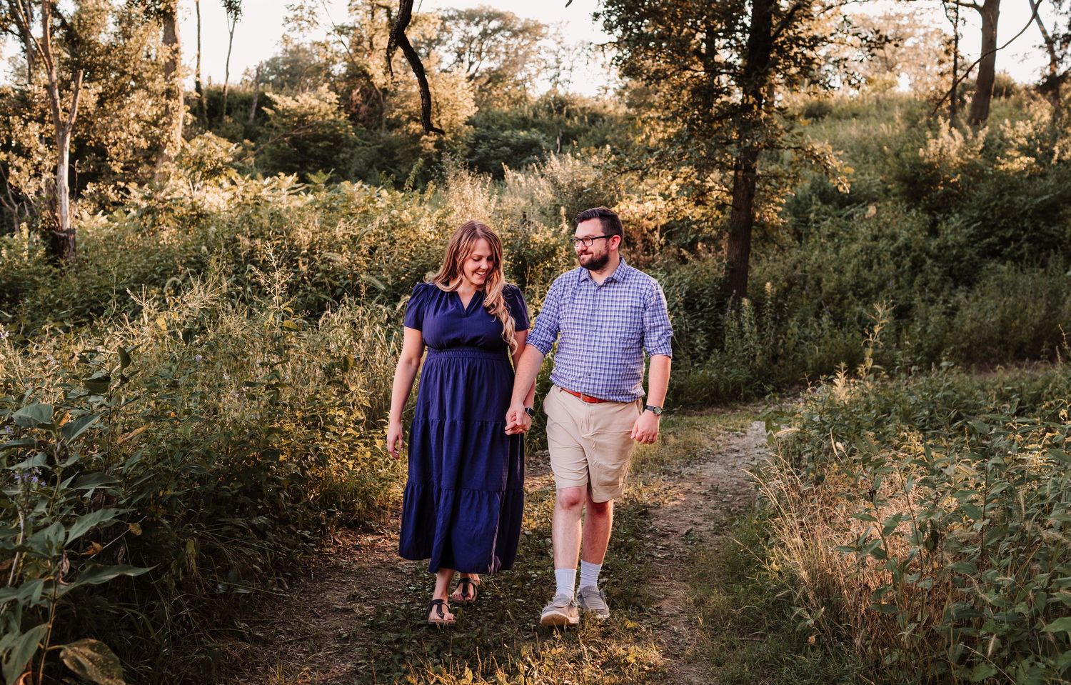 Couple holding hands walking on a nature trail surrounded by lush greenery during golden hour.